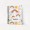 A spiral-bound notebook with a white cover decorated with colorful cartoon rainbows, smiling suns, clouds, stars, and a personalized name label reading 'Stephanie'. Blue dimension markers showing 6.