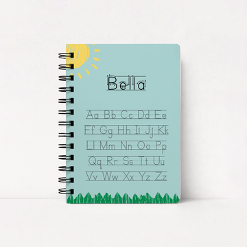 A personalized spiral-bound notebook with a light blue cover, featuring the name 'Bella' in cursive and the alphabet in lowercase letters, with a yellow sun and green grass illustration.