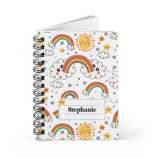 A spiral-bound notebook with a white cover decorated with cute cartoon rainbows, clouds, stars, and smiling suns. A white label on the front is printed with the name 'Stephanie' in black text.