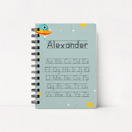 A spiral-bound notebook with a light blue cover featuring a cartoon alien, stars, and the name 'Alexander' above a dotted alphabet chart, shown on a clean white background.
