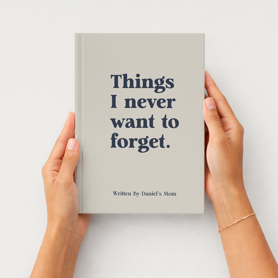 A person holds a hardcover book with both hands against a clean white background. The book cover is a warm light gray-beige with dark navy text that reads, 'Things I never want to forget.
