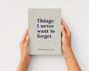 A person holds a hardcover book with both hands against a clean white background. The book cover is a warm light gray-beige with dark navy text that reads, 'Things I never want to forget.