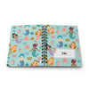 A personalized spiral-bound notebook with a light blue cover featuring a colorful mermaid pattern, shown open on a white background. The right page has a white label area printed with the name 'Ella'.