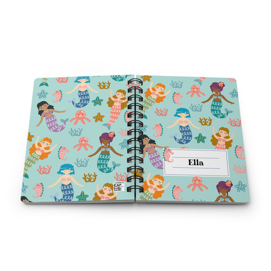 A personalized spiral-bound notebook with a light blue cover featuring a colorful mermaid pattern, shown open on a white background. The right page has a white label area printed with the name 'Ella'.