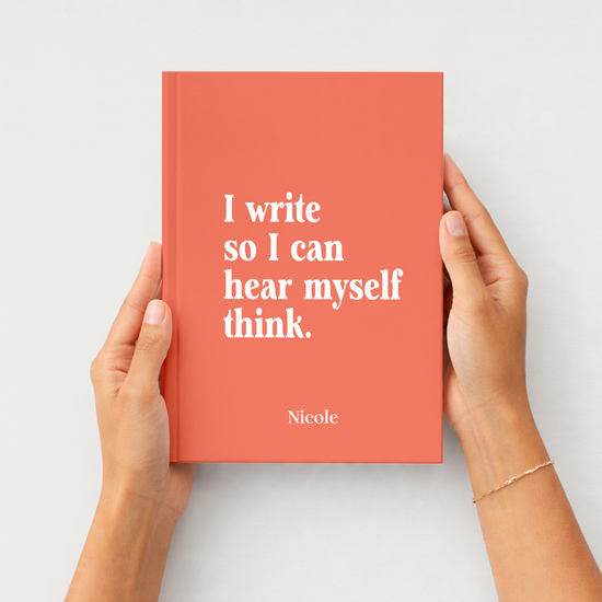 A coral red hardcover journal with white text reading 'I write so I can hear myself think.' and the name 'Nicole' at the bottom, held by two hands against a light gray background.
