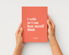 A coral red hardcover journal with white text reading 'I write so I can hear myself think.' and the name 'Nicole' at the bottom, held by two hands against a light gray background.
