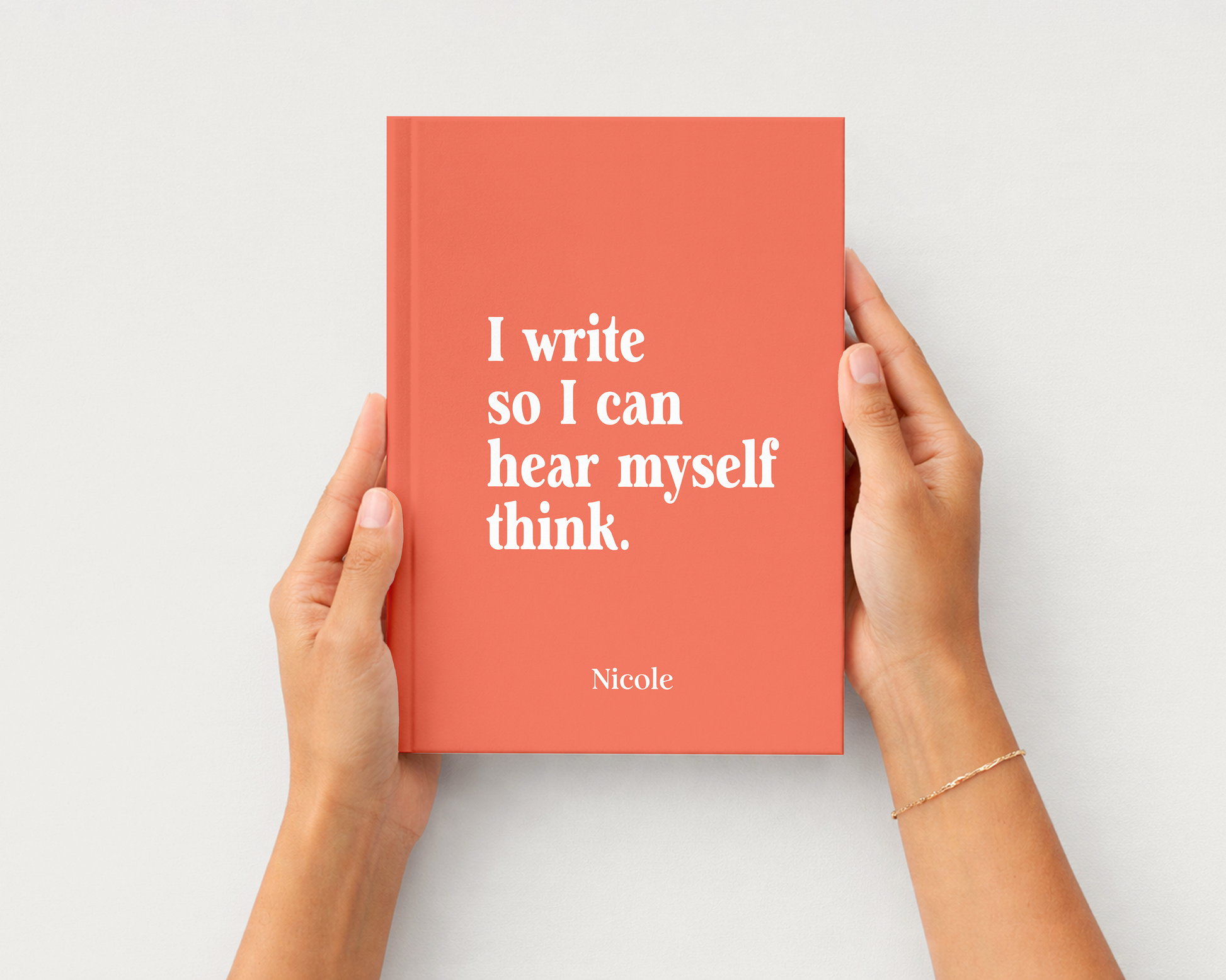 A coral red hardcover journal with white text reading 'I write so I can hear myself think.' and the name 'Nicole' at the bottom, held by two hands against a light gray background.