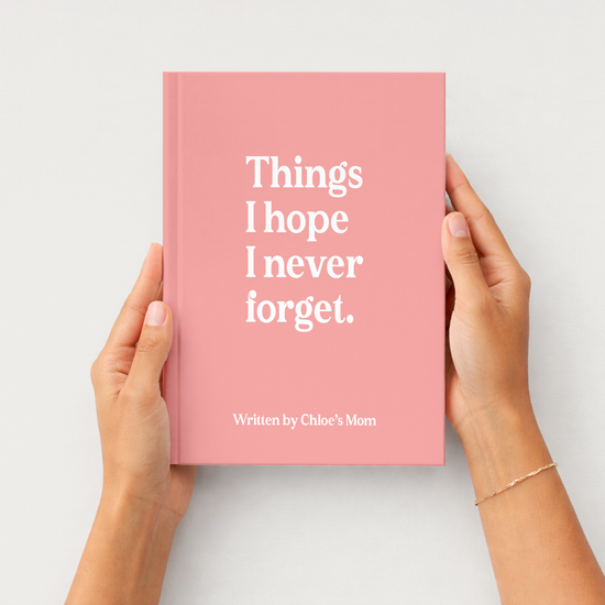 A pink hardcover book held by two hands against a light gray background. The book cover features the white text 'Things I hope I never forget.