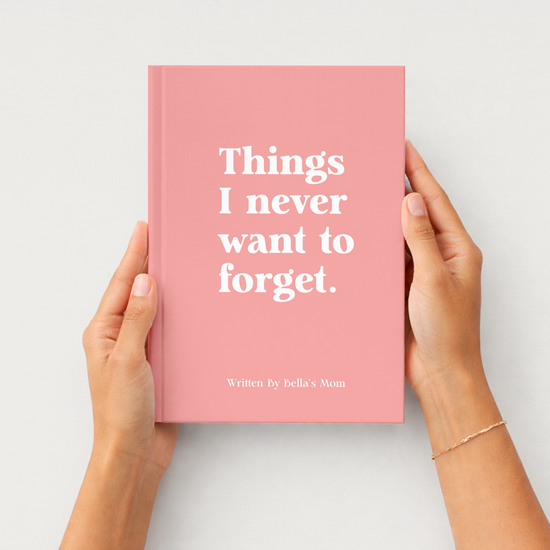 A pink hardcover notebook with white text reading 'Things I never want to forget. Written By: Jessica's Mom' is held by two hands against a clean white background.
