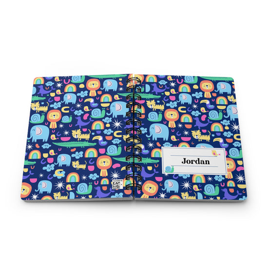 A personalized spiral-bound notebook with a colorful cartoon pattern of animals, rainbows, and stars on a dark blue background. The cover has a white label with the name 'Jordan' printed on it.
