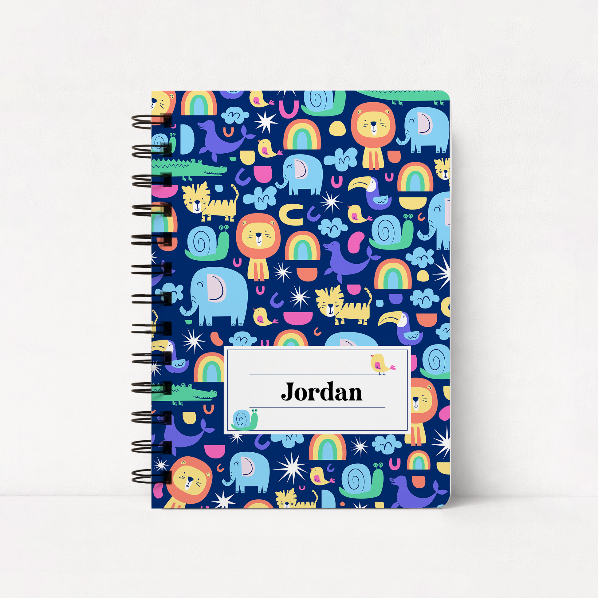 A spiral-bound notebook with a dark blue cover featuring a colorful cartoon safari pattern of lions, elephants, rainbows, and snakes. A white label on the front reads 'Jordan'.
