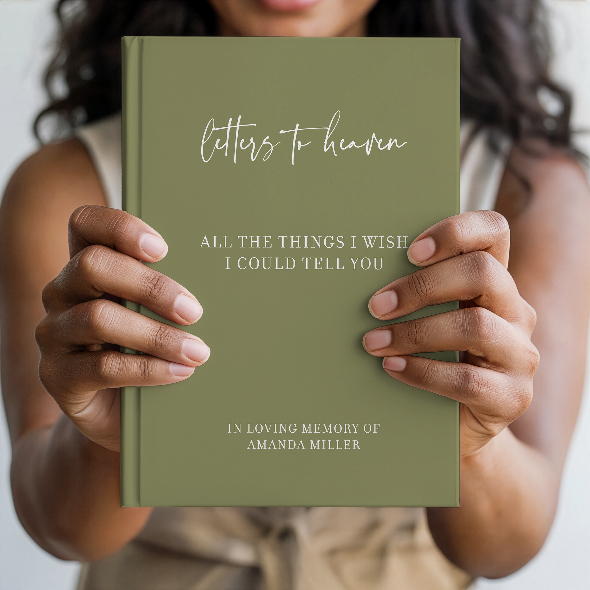 A person holds a hardcover book titled "Letters to heaven: All the things I wish I could tell you" in front of their face. The book cover is olive green with white text, and the background is a softly blurred beige-toned indoor setting.