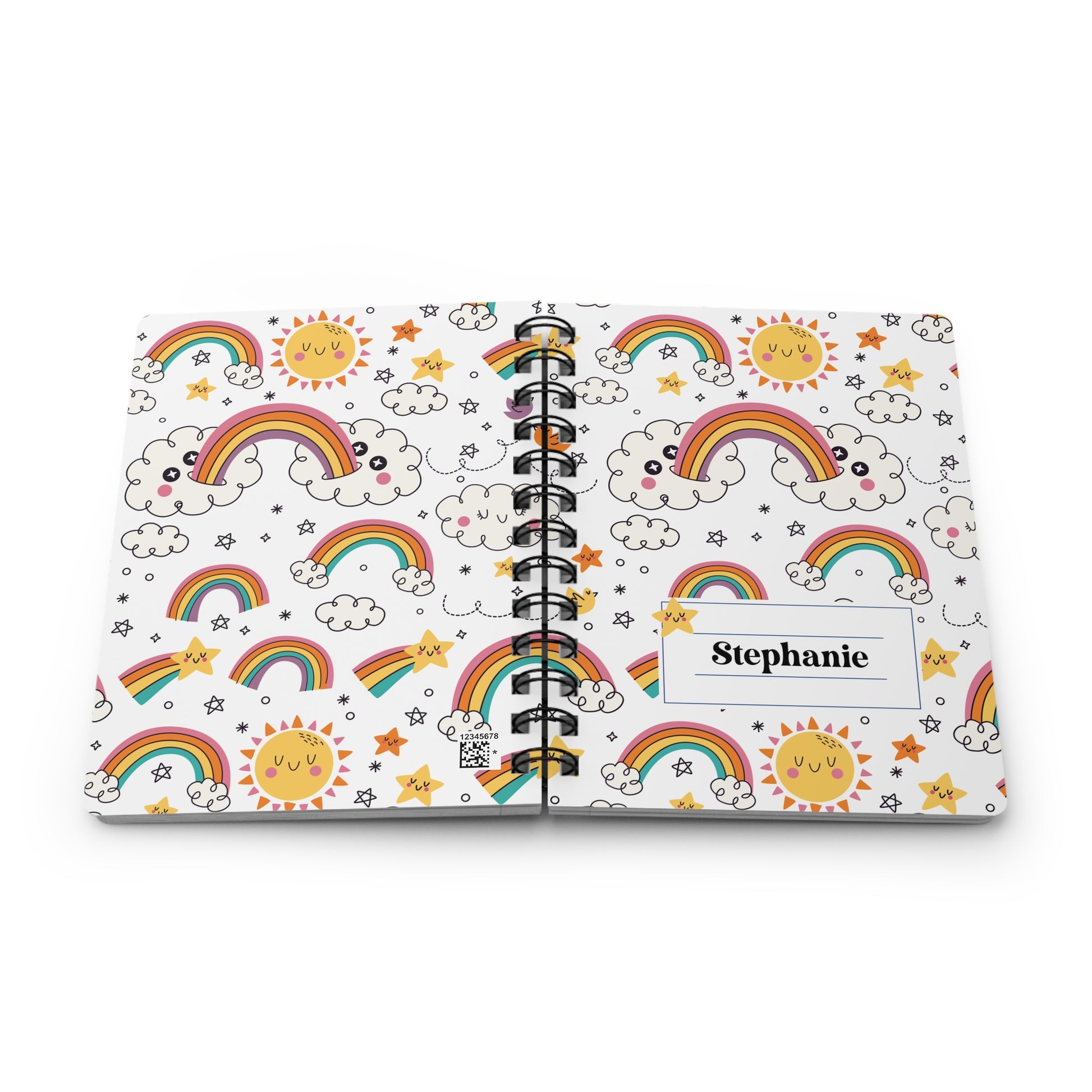 A personalized spiral-bound notebook with a white cover decorated with colorful cartoon rainbows, smiling suns, clouds, and stars. The name 'Stephanie' is printed in black text inside a white label on the front cover.