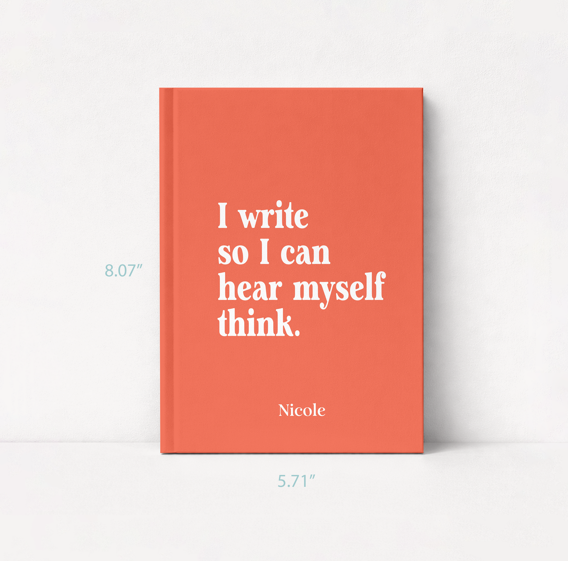 A coral orange hardcover book with white text reading 'I write so I can hear myself think.' and the name 'Nicole' at the bottom, shown standing against a white wall.