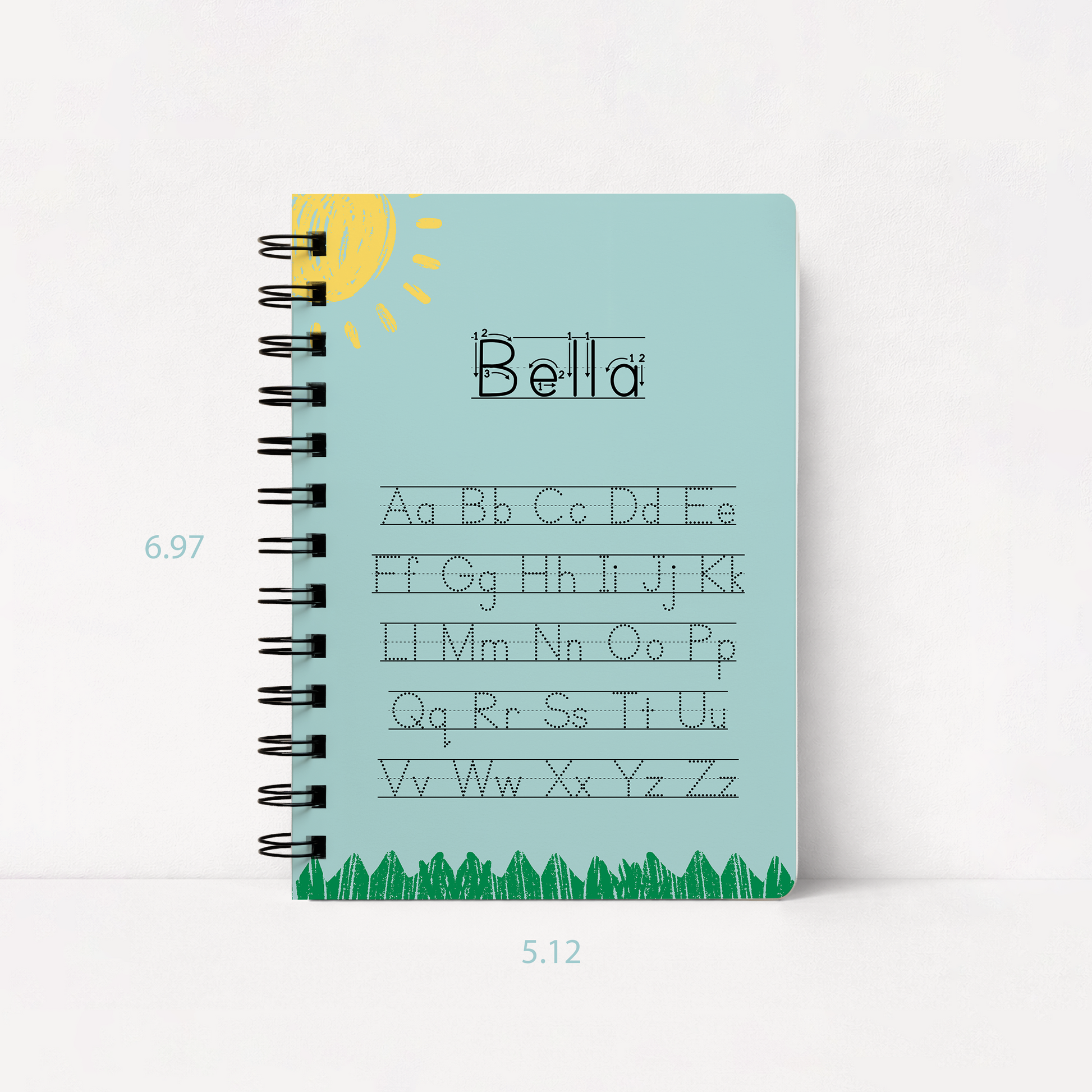 A spiral-bound notebook with a light blue cover, featuring the word 'Bella' in outlined letters, alphabet practice text, a yellow sun illustration, and green grass along the bottom. The notebook is shown on a white background with dimension labels indicating 6.97.