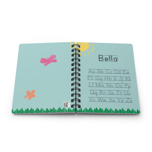 A personalized alphabet journal for 'Bella' with a light blue cover, featuring a black spiral binding, a yellow sun, pink and orange butterflies, green grass, and printed alphabet letters from A to Z.