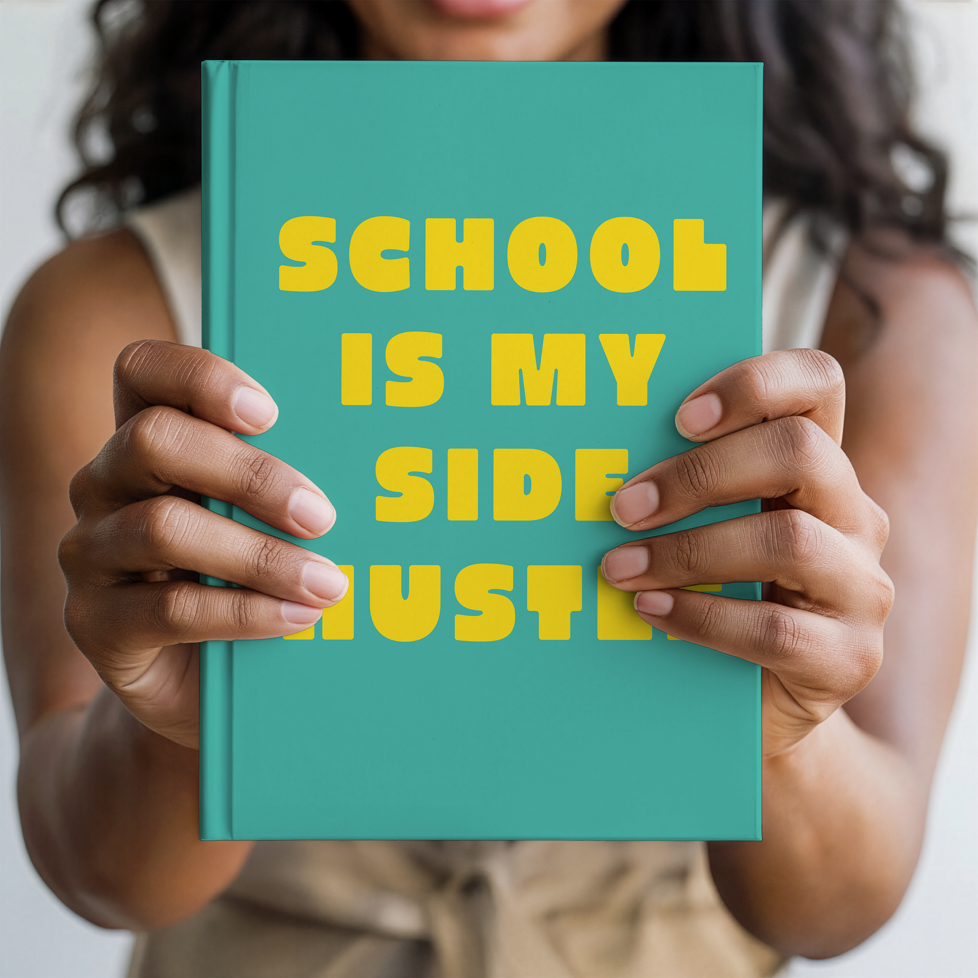 A person holding a teal notebook with bold yellow text that reads 'SCHOOL IS MY SIDE HUSTER' against a soft, blurred background.