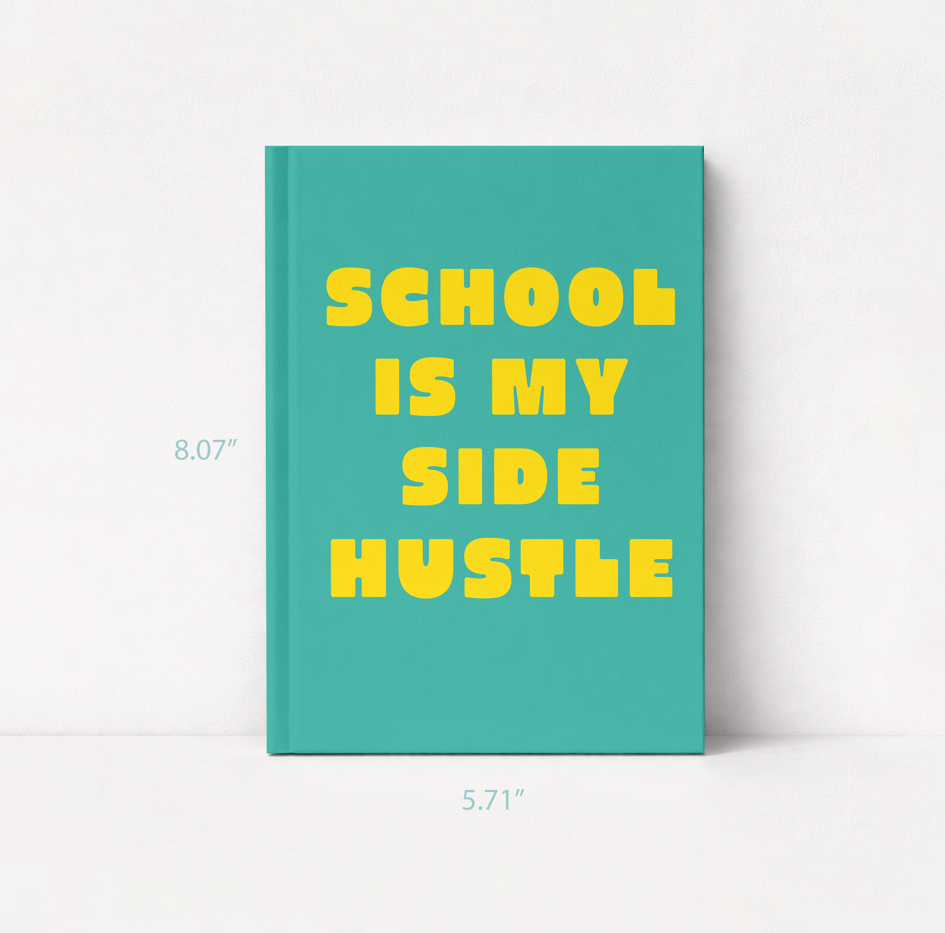 A teal hardcover notebook or journal standing upright on a white surface against a white wall, with large yellow text reading “SCHOOL IS MY SIDE HUSTLE.” 