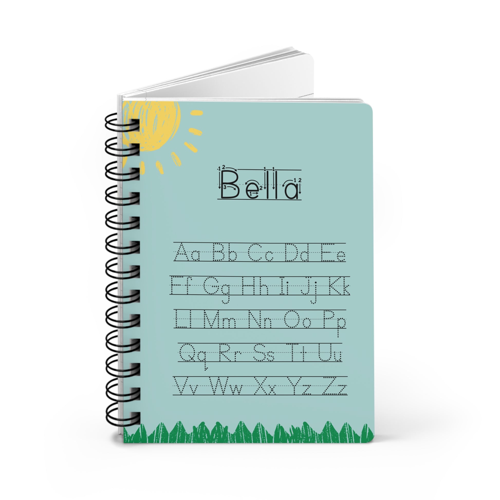 A spiral-bound notebook with a light blue cover featuring the word 'Bella' in cursive, alphabet practice letters, a yellow sun illustration, and green grass along the bottom, shown on a white background.