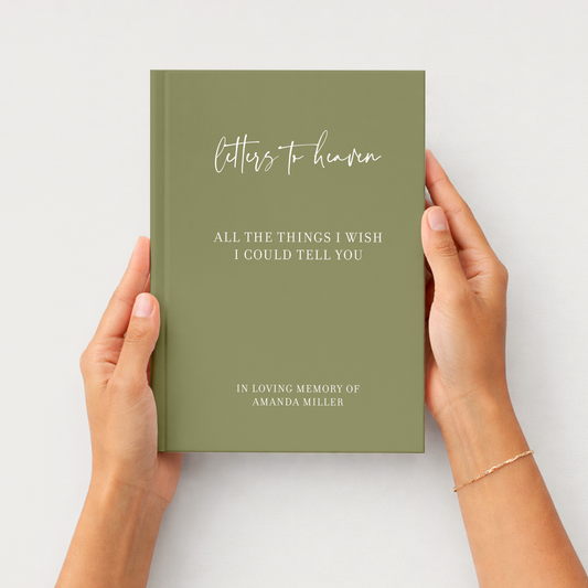 A person holds a hardcover journal with an olive green cover. The cover features white text reading 'letters to heaven' and 'ALL THE THINGS I WISH I COULD TELL YOU' with 'IN LOVING MEMORY OF AMANDA MILLER' at the bottom.